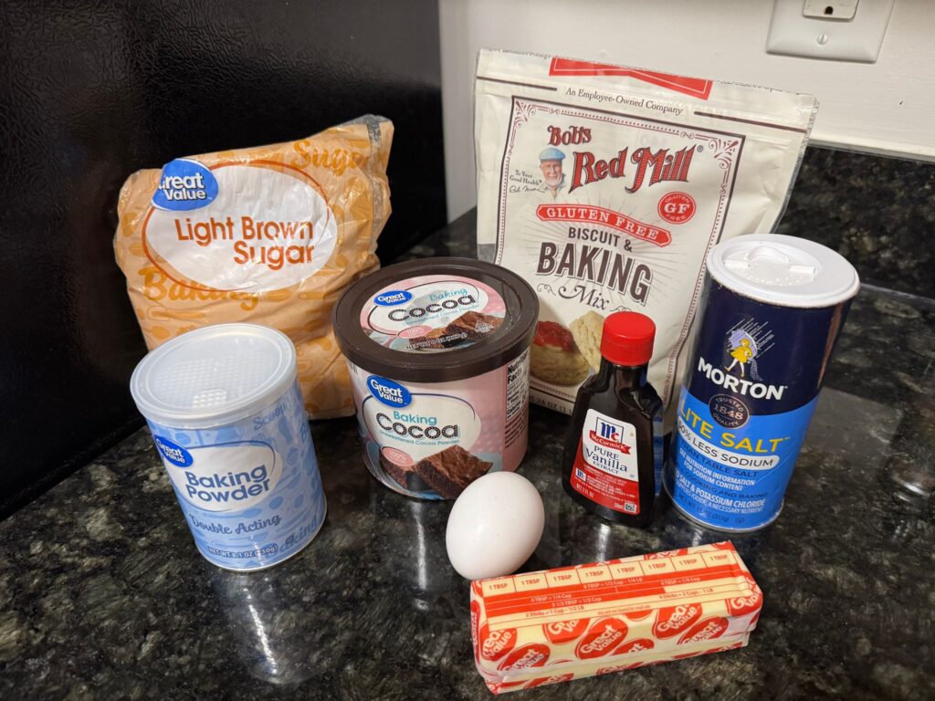 This picture shows the ingredients used to make the chocolate crinkle cookies. Gluten free baking mix, brown sugar, baking powder, cocoa powder, egg, butter, vanilla, salt, and powdered sugar.