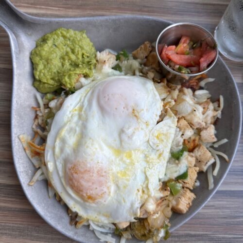 This is a picture of a breakfast skillet. It has chicken, grilled bell peppers, cheese, hash browns, topped with 2 over-easy eggs. On the side there is guacamole and salsa.