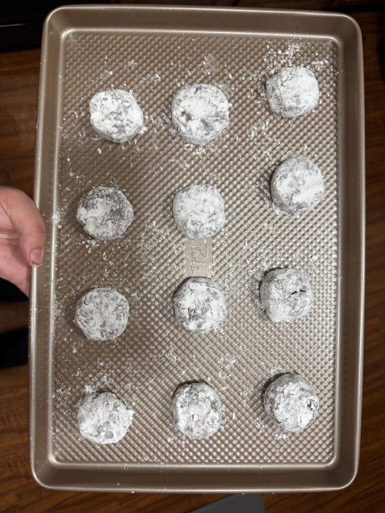 This pictures shows 12 crinkle cookies equally spaced out on a cookie sheet, coated in powdered sugar.