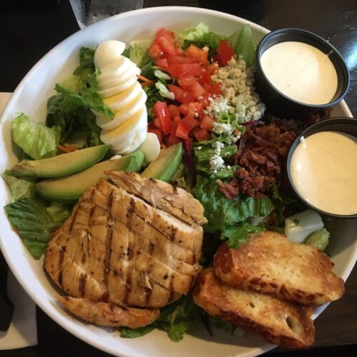 This image is of a cobb salad. It has romaine lettuce topped with grilled chicken, egg, avocado, bacon, tomatoes and salad dressing.